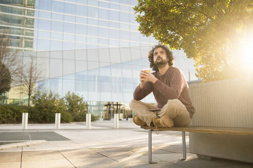 Portrait of bearded man relaxing on bench with coffee to go at sunset - KIJF02974