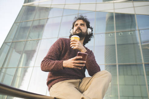Portrait of bearded man sitting outdoors with coffee to go, headphones and smartphone - KIJF02977