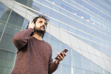 Portrait of bearded man with headphones and smartphone outdoors looking at distance - KIJF02978