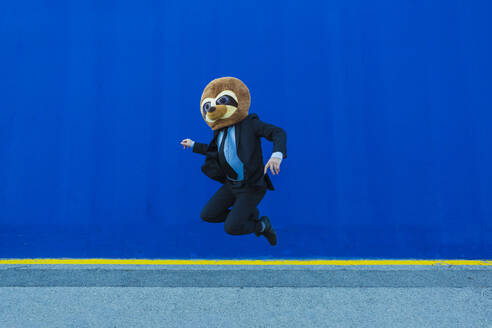 Geschäftsmann im schwarzen Anzug mit Erdmännchen-Maske, der vor einer blauen Wand in die Luft springt - XLGF00037