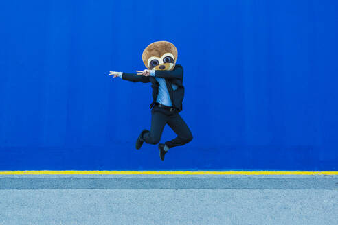 Businessman in black suit with meerkat mask jumping in the air in front of blue wall - XLGF00039