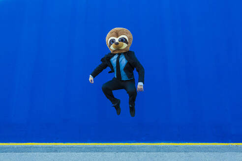Geschäftsmann im schwarzen Anzug mit Erdmännchen-Maske, der vor einer blauen Wand in die Luft springt - XLGF00042