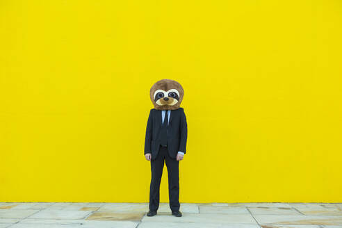 Geschäftsmann im schwarzen Anzug mit Erdmännchen-Maske vor gelber Wand stehend - XLGF00045