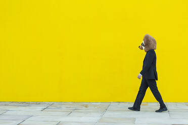 Businessman in black suit with meerkat mask walking in front of yellow wall - XLGF00055