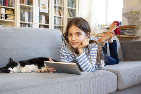 Portrait of smiling girl lying on couch with cat and digital tablet - LVF08825