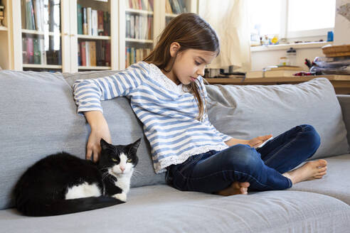 Mädchen sitzt auf der Couch und benutzt ein digitales Tablet, während sie ihre Katze streichelt - LVF08826