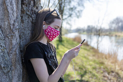 Girl with homemade protective mask using smartphone and leaning on tree - SARF04536