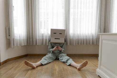 Woman wearing a cardbox on head with bored smiley sitting on ground and using smartphone - AFVF06051