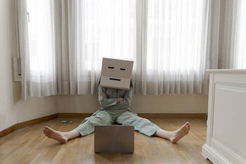 Woman wearing a cardbox on head with bored smiley sitting on floor and looking at laptop - AFVF06054