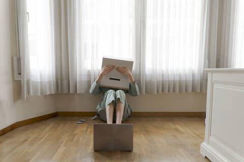 Woman wearing a cardbox on head with bored smiley sitting on floor in front of laptop, covering the eyes stock photo