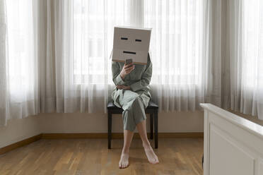 Woman wearing a cardbox on head with bored smiley sitting on chair and using smartphone - AFVF06058