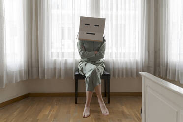 Frau, die einen Karton auf dem Kopf mit einem gelangweilten Smiley trägt und mit verschränkten Armen auf einem Stuhl sitzt - AFVF06059