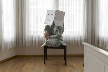 Frau mit Karton auf dem Kopf und gelangweiltem Smiley sitzt auf einem Stuhl - AFVF06060