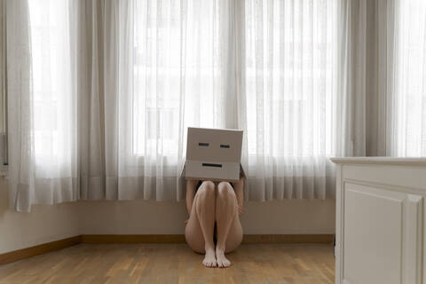 Frau trägt einen Karton auf dem Kopf mit einem gelangweilten Smiley und sitzt auf dem Boden, lizenzfreies Stockfoto