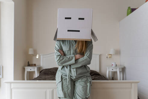 Woman wearing a cardbox on head with bored smiley leaning on bed at home - AFVF06067