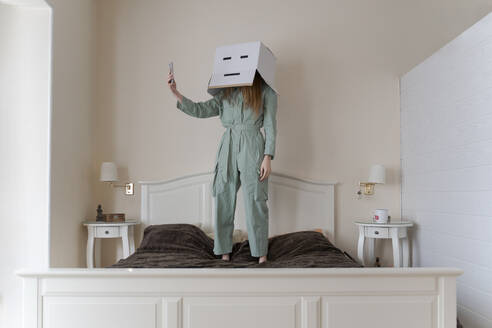 Woman wearing a cardbox on head with bored smiley taking a selfie on her bed - AFVF06068