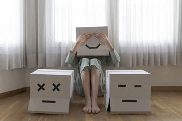 Woman wearing a cardbox on head with bored smiley sitting on floor in front of laptop, covering the eyes - AFVF06076