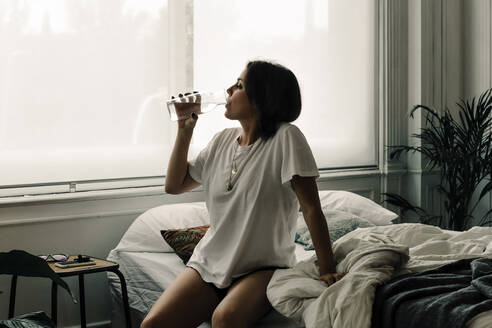 Mature woman sitting on bed in the morning drinking water - ERRF03473