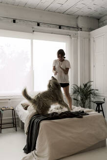 Mature woman standing on bed playing with her dog - ERRF03477