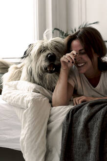 Laughing mature woman lying on bed playing with her dog - ERRF03483