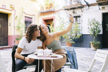 Junge Frau, die mit ihrem Freund ein Selfie mit dem Handy macht, während sie in einem Straßencafé sitzt, Santa Cruz, Sevilla, Spanien - DGOF00856