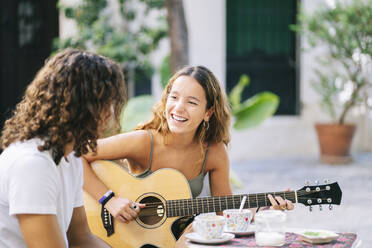 Glückliche junge Frau spielt Gitarre, während sie mit ihrem Freund in einem Straßencafé sitzt, Santa Cruz, Sevilla, Spanien - DGOF00859