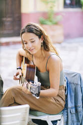 Smiling young woman playing guitar while sitting at sidewalk cafe, Santa Cruz, Seville, Spain - DGOF00860