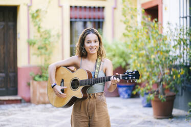 Porträt einer lächelnden jungen Frau, die Gitarre spielt, während sie auf der Straße steht, Santa Cruz, Sevilla, Spanien - DGOF00861