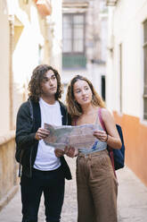 Junges Paar, das auf einer schmalen Straße in Santa Cruz, Sevilla, Spanien, mit einer Karte in der Hand wegschaut - DGOF00864