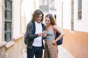 Smiling young woman looking at man using smart phone while standing on narrow street at Santa Cruz, Seville, Spain - DGOF00866