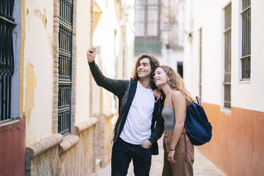 Lächelndes junges Paar, das ein Selfie macht, während es in einer engen Straße in Santa Cruz, Sevilla, Spanien steht - DGOF00867