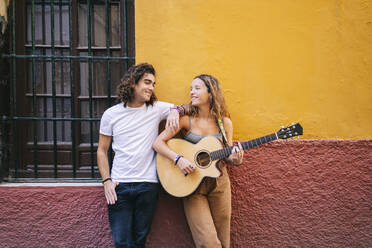 Lächelnder junger Mann, der neben seiner Freundin steht und Gitarre spielt, während er an der Wand steht, Santa Cruz, Sevilla, Spanien - DGOF00870