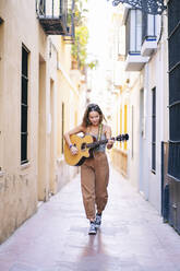 Junge Frau in voller Länge beim Gitarrenspiel in einer engen Straße inmitten von Gebäuden in Santa Cruz, Sevilla, Spanien - DGOF00874