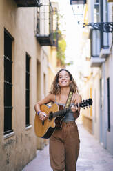 Porträt einer lächelnden jungen Frau, die Gitarre spielt, während sie durch eine enge Straße in Santa Cruz, Sevilla, Spanien geht - DGOF00875