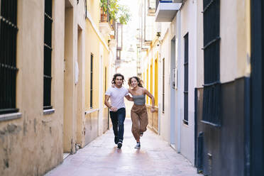 Unbeschwertes junges Paar läuft in voller Länge auf einer engen Straße in Santa Cruz, Sevilla, Spanien - DGOF00878