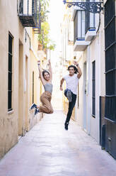 Unbeschwertes Paar in voller Länge beim Springen auf einer schmalen Straße während des Urlaubs in Santa Cruz, Sevilla, Spanien - DGOF00880