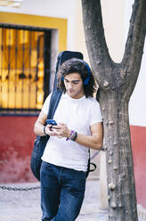 Junger Mann mit Mobiltelefon und Gitarrentasche, Santa Cruz, Sevilla, Spanien - DGOF00882