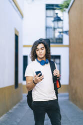 Junger Mann, der ein Mobiltelefon benutzt, während er in einer engen Straße in Santa Cruz, Sevilla, Spanien, steht - DGOF00883