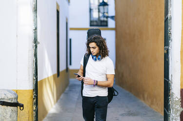 Junger Mann, der ein Mobiltelefon benutzt, während er durch eine enge Straße in Santa Cruz, Sevilla, Spanien, geht - DGOF00884