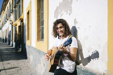 Lächelnder junger Mann spielt Gitarre und lehnt sich an eine Wand, Santa Cruz, Sevilla, Spanien - DGOF00885