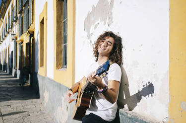 Lächelnder junger Mann mit geschlossenen Augen, der Gitarre spielt und sich an eine Wand in Santa Cruz, Sevilla, Spanien, lehnt - DGOF00886