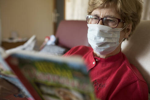 Senior woman reading roman at home and wearing protective mask - JATF01179