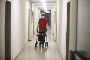 Senior woman wearing mask and walking with wheeled walker in corridor of retirement home - JATF01181
