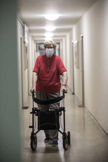 Senior woman wearing mask and walking with wheeled walker in corridor of retirement home - JATF01182