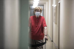 Senior woman wearing mask and walking with wheeled walker in corridor of retirement home - JATF01183