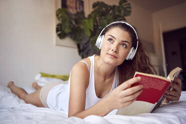 Porträt einer jungen Frau mit Buch, die auf dem Bett liegt und mit Kopfhörern Musik hört - JHAF00103