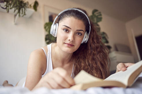 Portrait of young woman with book lying on bed listening music with headphones - JHAF00104