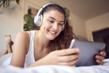 Porträt einer lächelnden jungen Frau, die mit einem digitalen Tablet auf dem Bett liegt und mit Kopfhörern Musik hört - JHAF00105