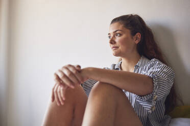 Porträt einer jungen Frau, die auf einem Bett sitzt und in die Ferne schaut - JHAF00107