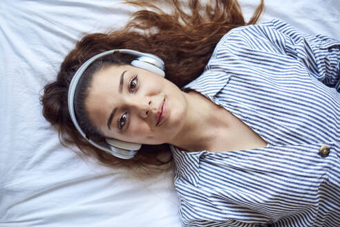 Porträt einer lächelnden jungen Frau, die auf dem Bett liegt und mit Kopfhörern Musik hört - JHAF00109